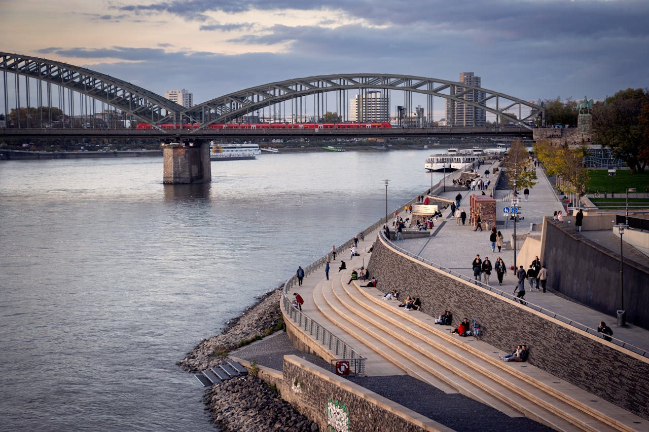 A picturesque urban scene in Cologne, Germany with Hohenzollern Bridge, Rhine River, and people enjoying the riverside.